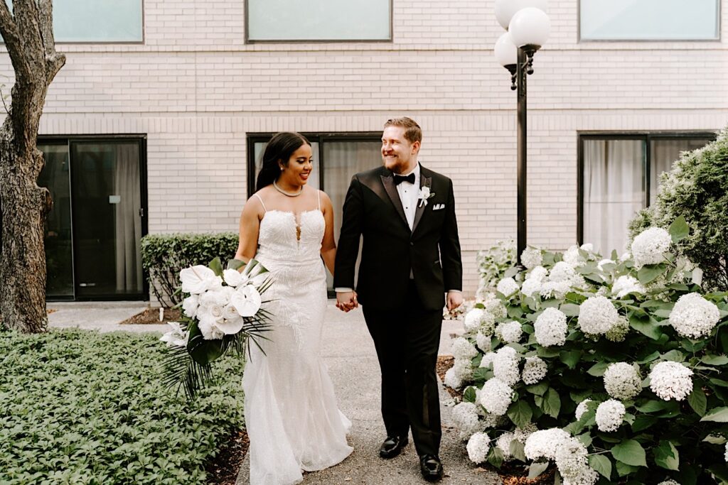 A bride and groom hold hands and walk on a sidewalk in a courtyard towards the camera while smiling at one another