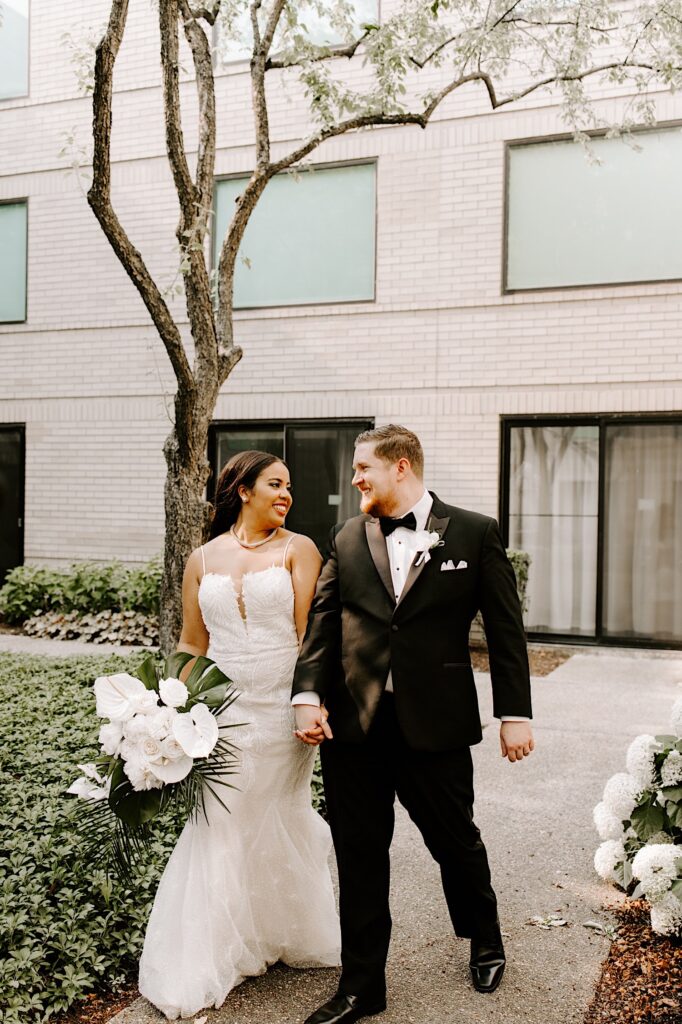 A bride and groom smile at one another as they walk along an outdoor path in a courtyard