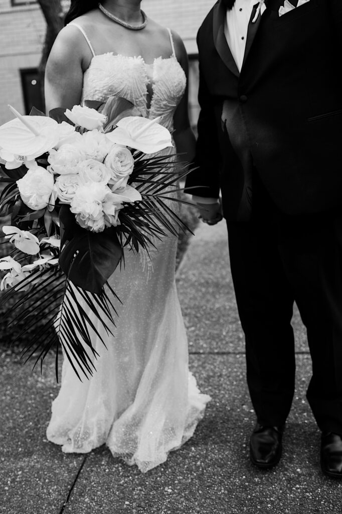 Black and white photo of a bride and groom standing side by side and holding hands while the bride holds her white floral bouquet