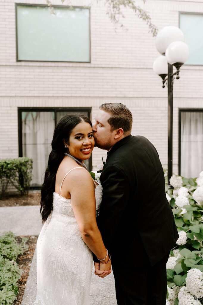 A bride smiles over her shoulder at the camera as the groom kisses her on the cheek while the two stand outside in a courtyard