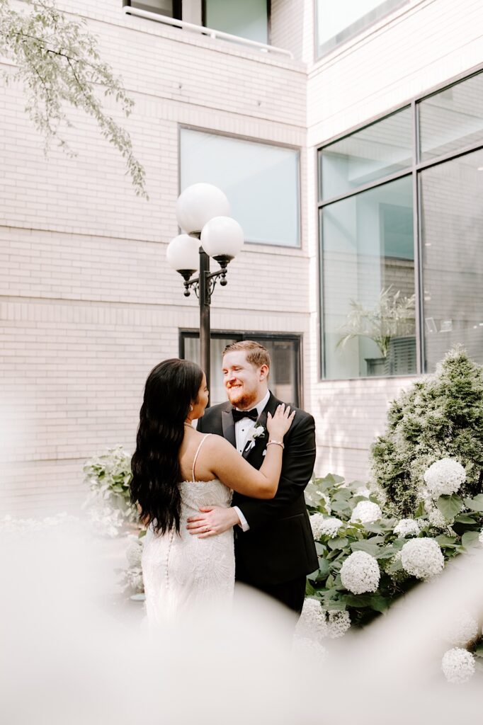 A groom smiles at the bride as the two embrace while standing in a courtyard