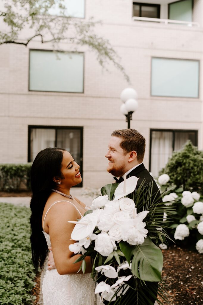 A bride and groom smile at each other while standing outside in a courtyard together