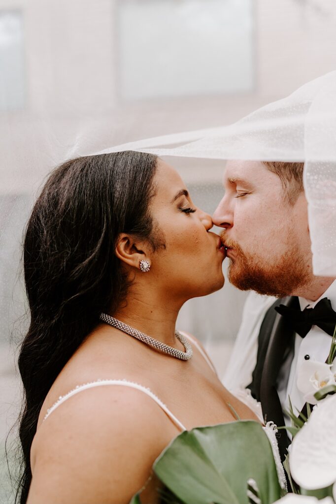 A bride and groom kiss one another while standing underneath the bride's veil