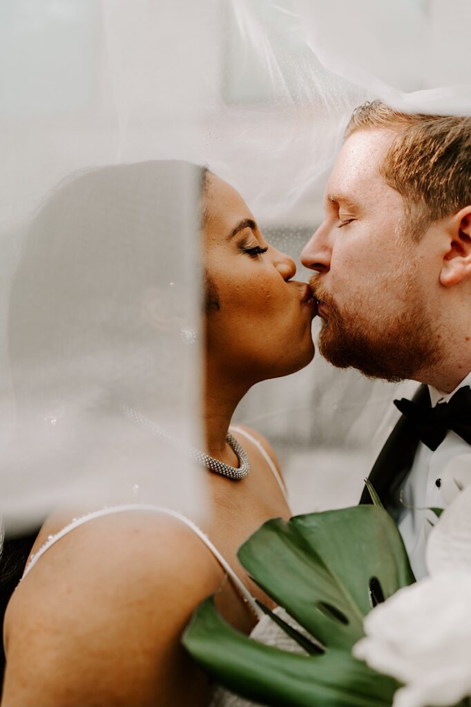 A bride and groom kiss one another while standing underneath the bride's veil