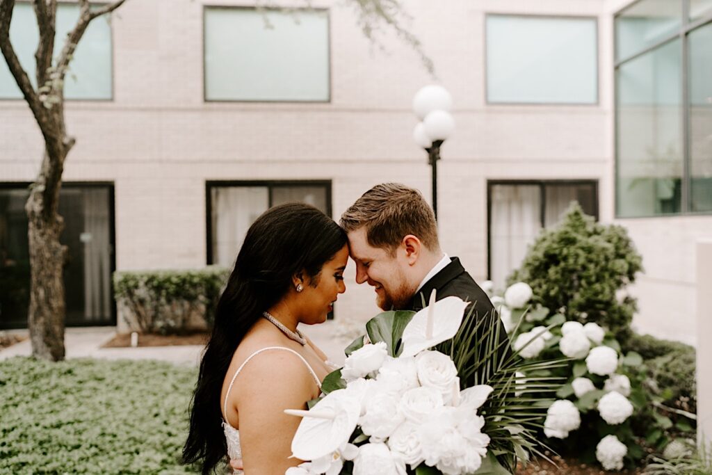 A bride and groom smile as they touch foreheads together and smile while standing together in an outdoor courtyard