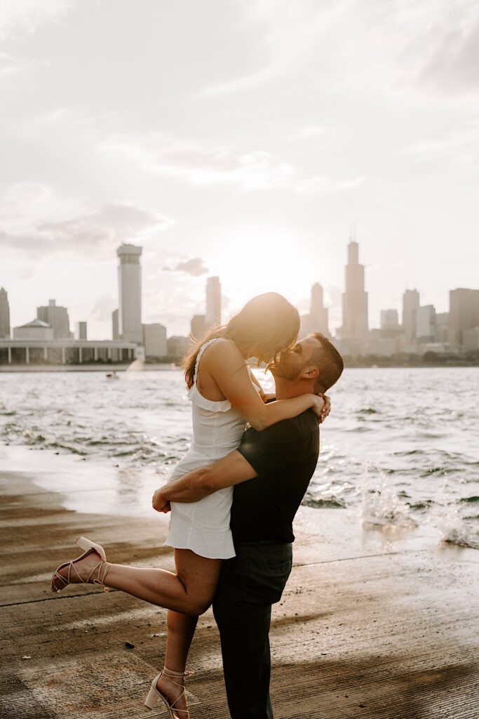 A man lifts a woman in into the air and kisses her while standing next to Lake Michigan with the sunset and the Chicago skyline behind them