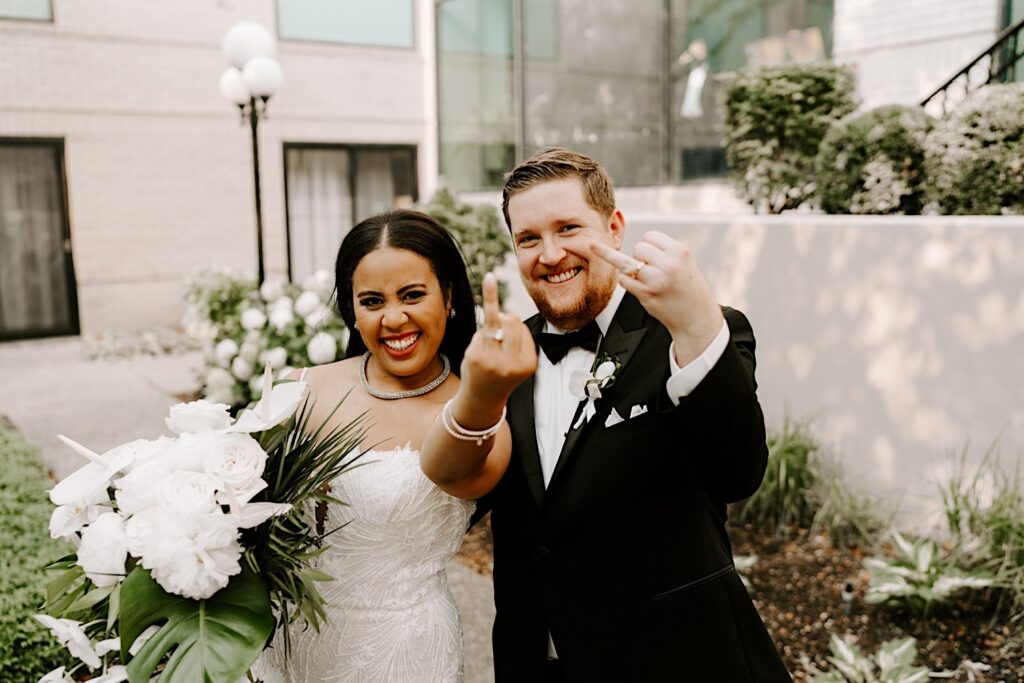 A bride and groom smile while showing off their ring fingers to the camera on their wedding day, the two are standing outside of a building with different plants around them