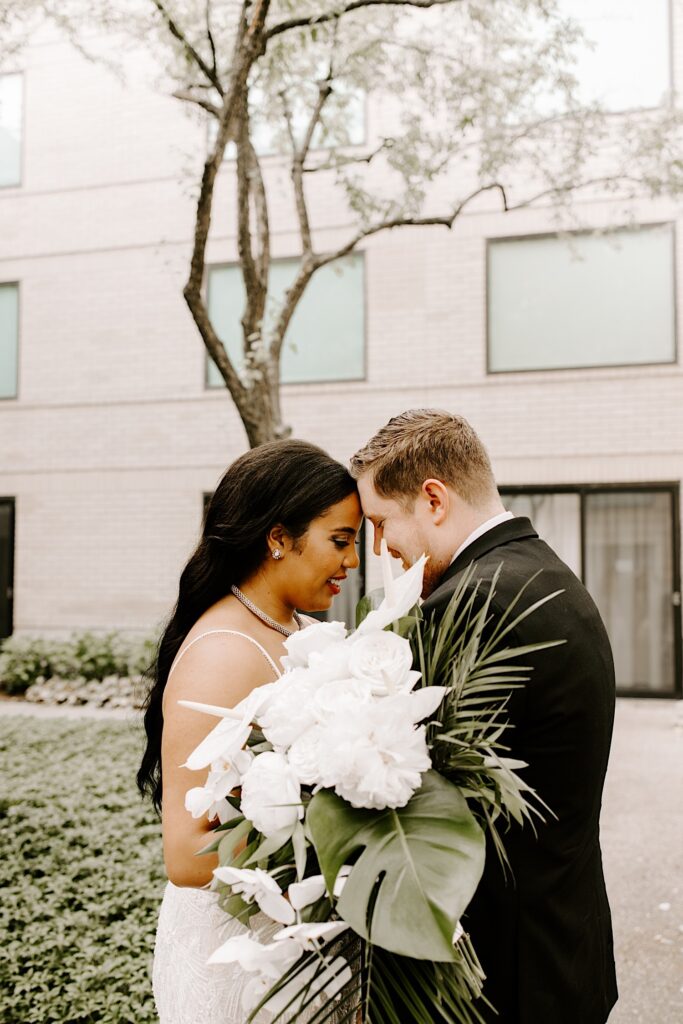 A bride and groom smile while touching foreheads together while standing outside in a courtyard