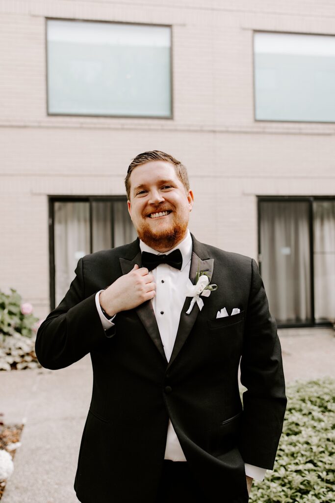 Portrait photo of a groom smiling at the camera and adjusting his suit coat while standing outside in a courtyard
