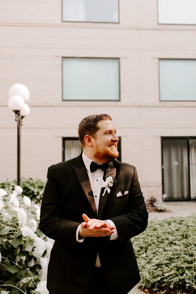 Portrait of a groom smiling to the right while rubbing his hands together as he stands outside in a courtyard