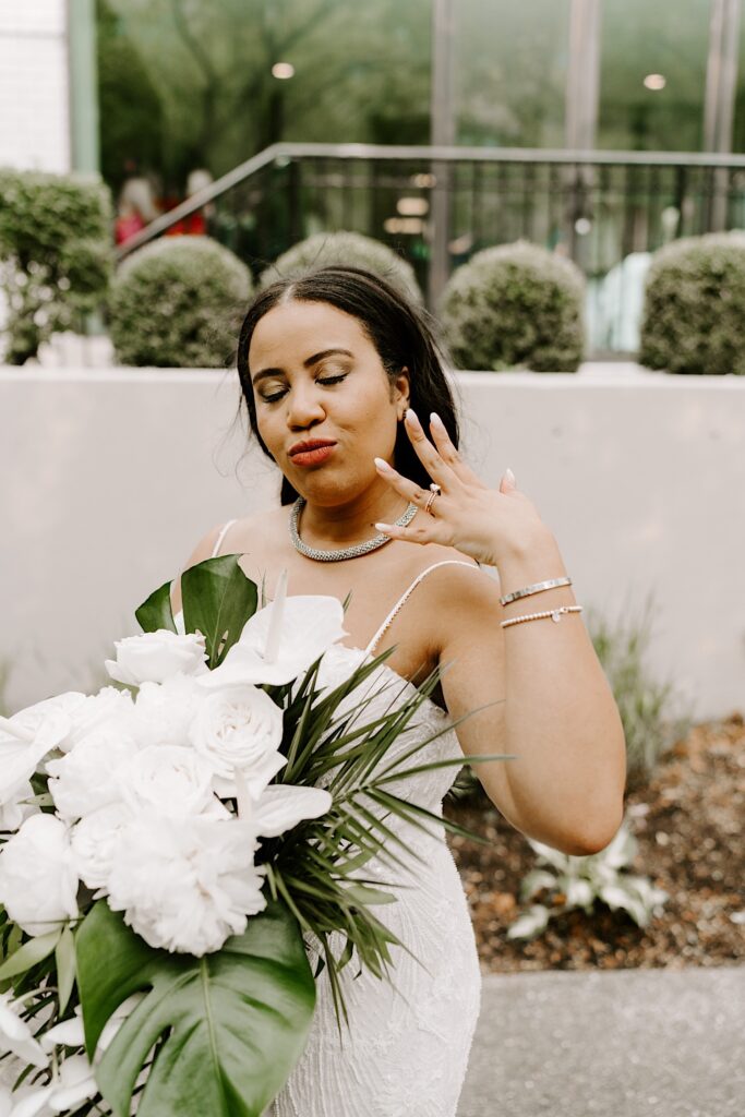 Portrait photo of a bride kissing towards the camera with her eyes closed as she shows off her wedding ring while standing outside