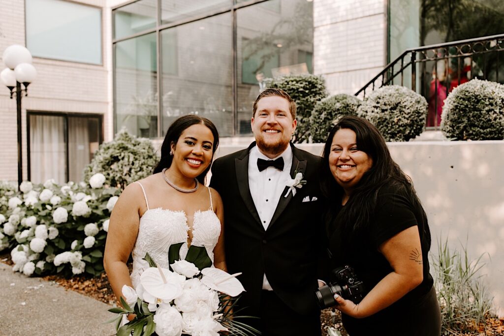 A bride and groom smile at the camera alongside their photographer JNA Visuals who is also smiling at the camera, the three are standing outdoors in a courtyard