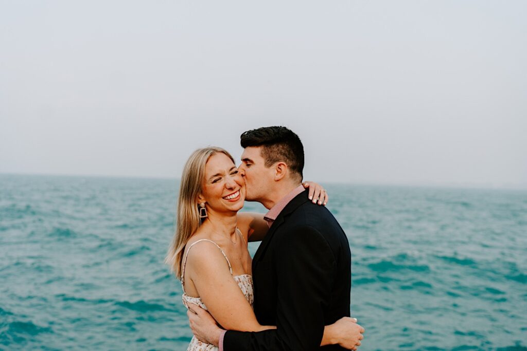 A woman smiles with her eyes closed while hugging a man who kisses her on the cheek in front of Lake Michigan