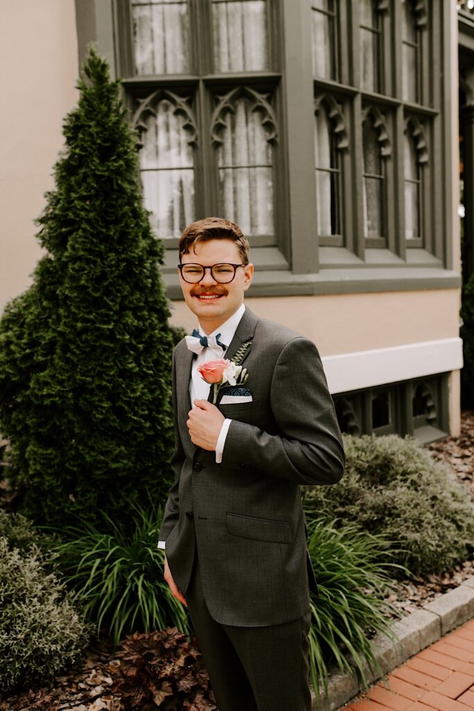 Portrait photo of a groom smiling at the camera while posing in front of a building with a tree and bushes behind him