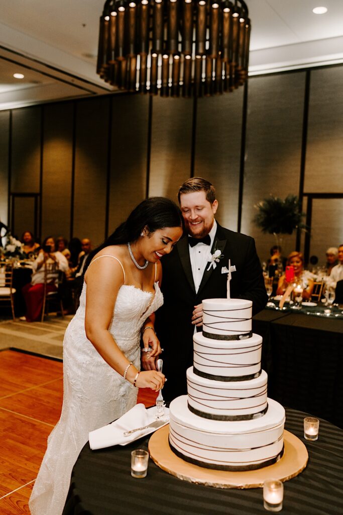 A bride and groom smile as they cut their wedding cake together during their indoor wedding reception