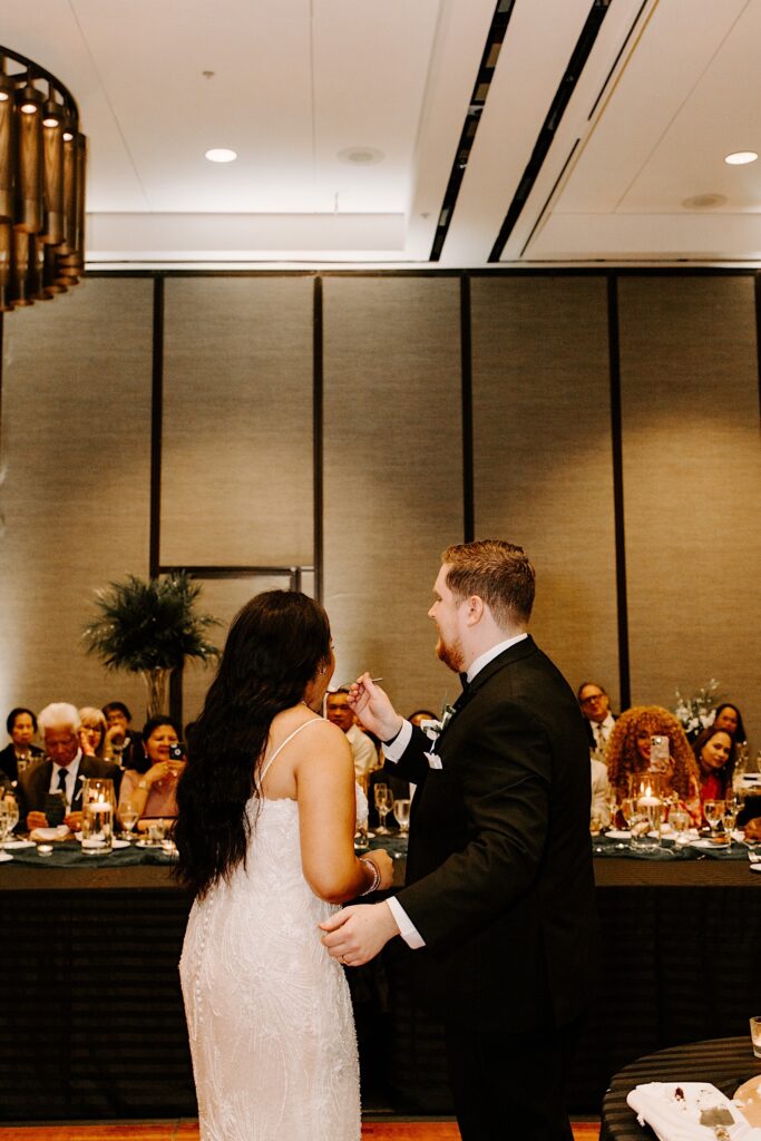 A bride is fed a piece of cake by the groom during their indoor wedding reception