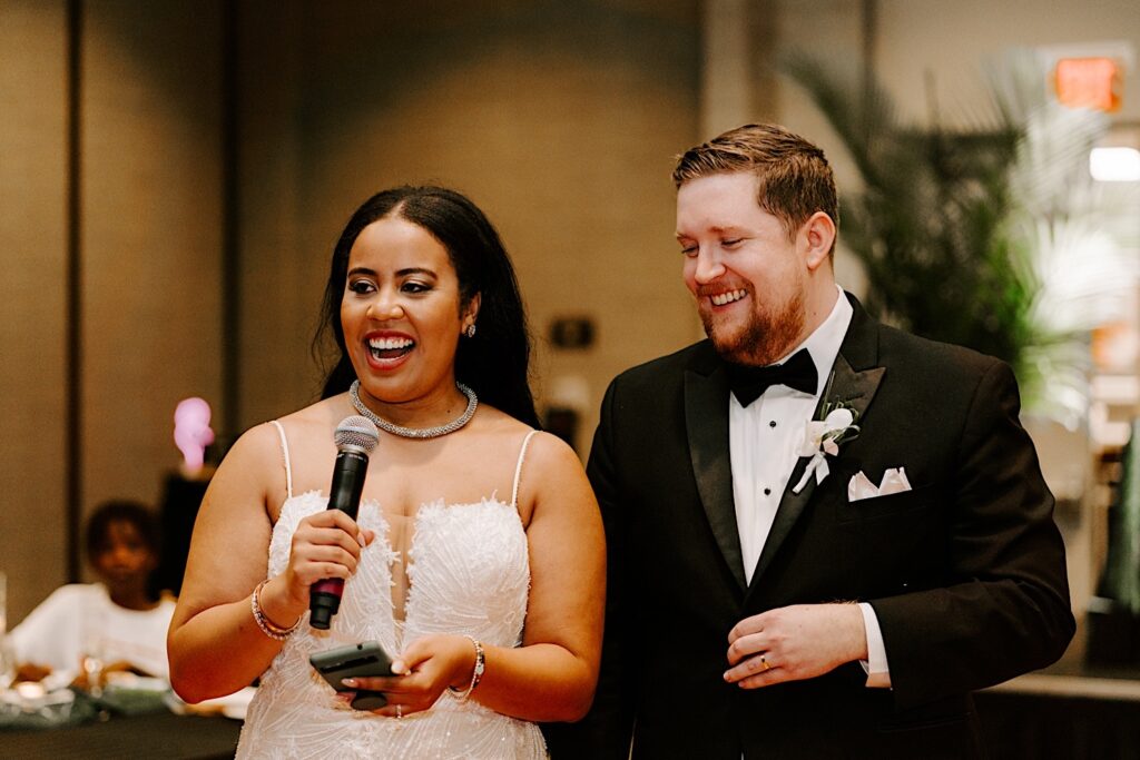A bride speaks while holding a microphone as the groom smiles at her side during their indoor wedding reception at the Hyatt Regency