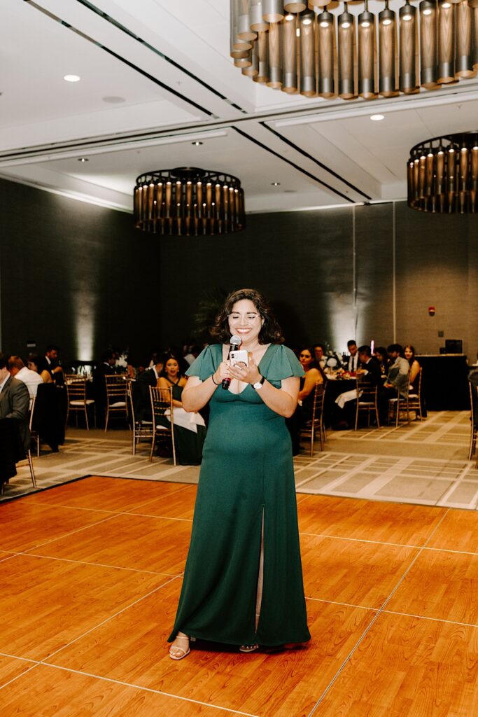 A bridesmaid stands in the middle of a dancefloor while holding a microphone to give a speech during an indoor wedding reception