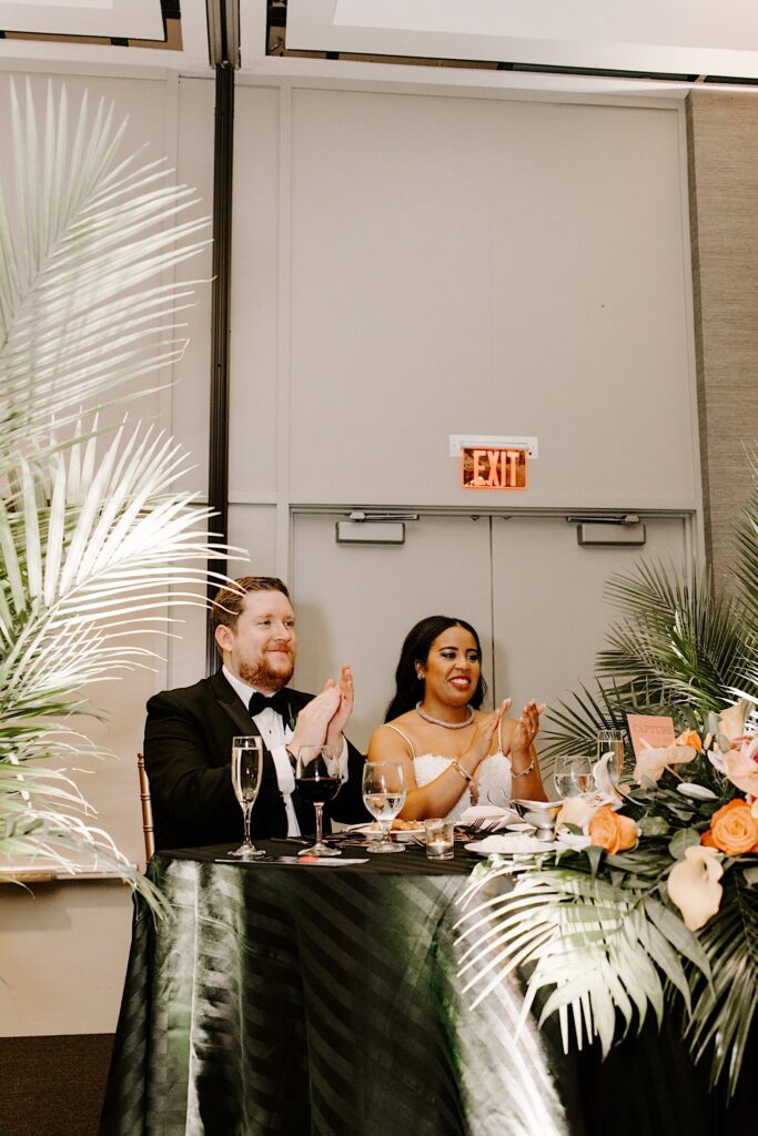 A bride and groom sit together at a table and clap during a speech at their indoor wedding reception
