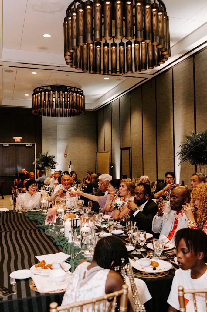 Guests of a wedding reception sit at a table together and raise their glasses during a speech