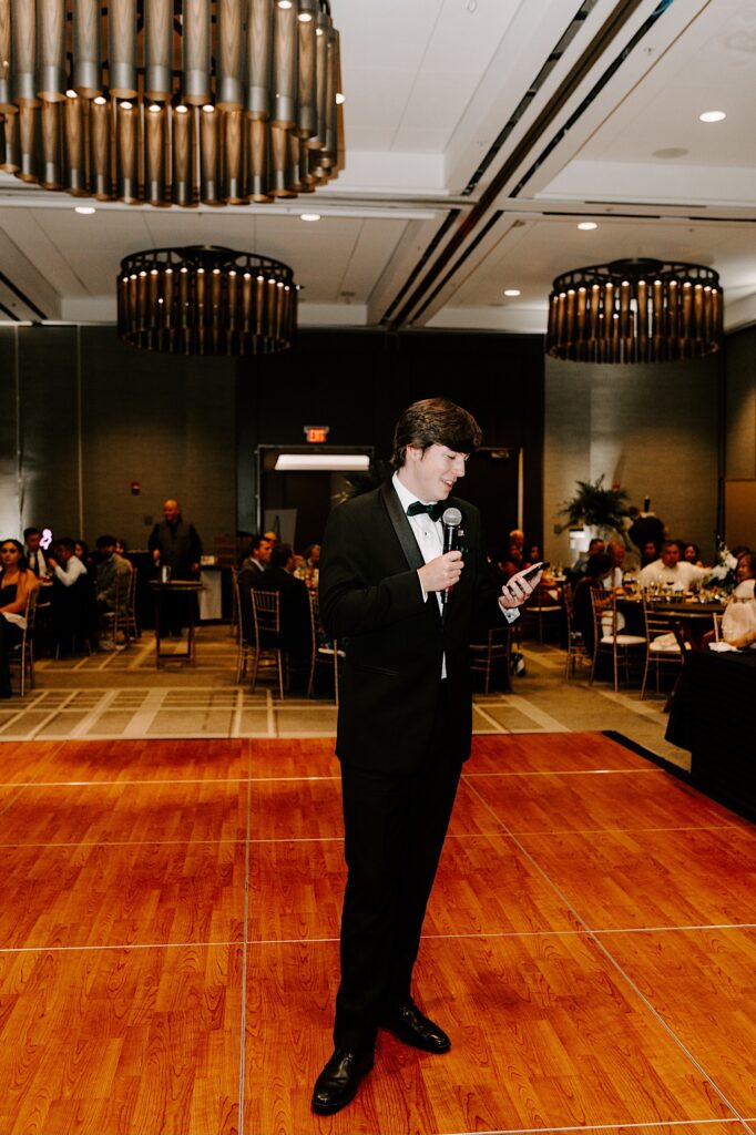 A groomsman stands in the middle of a dancefloor and speaks into a microphone during an indoor wedding reception