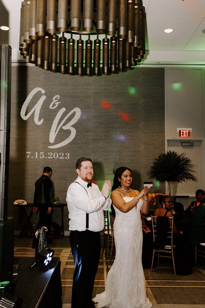 A bride and groom clap and smile while standing off to the side of the dance floor of their wedding reception