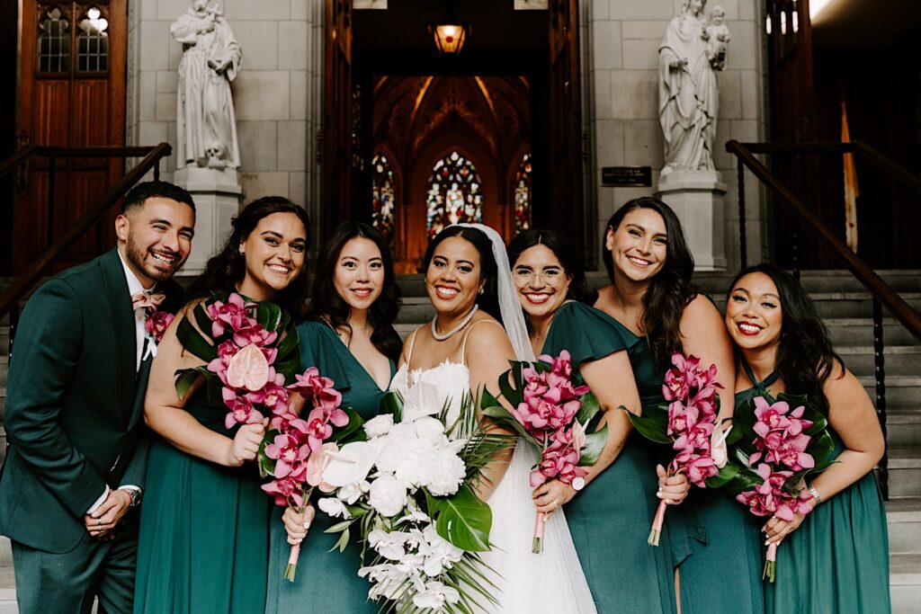 A bride and her wedding party stand next to one another while outside of a church and smile at the camera while holding their flower bouquets