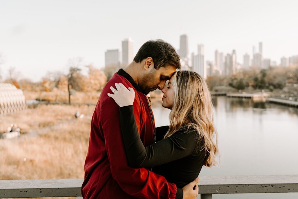 A man and woman touch their foreheads together and embrace with their eyes closed while standing in Lincoln Park during their engagement photos, behind them is the Chicago skyline