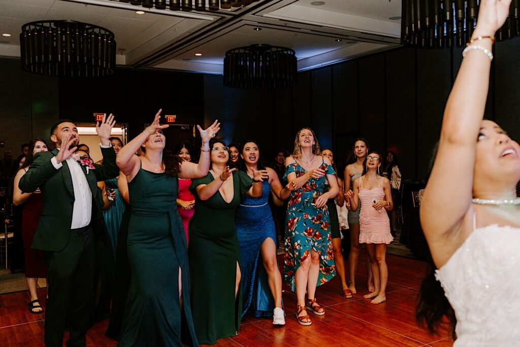Guests of an indoor wedding reception at the Hyatt Regency look up and raise their arms to catch the bouquet that the bride just threw in the air