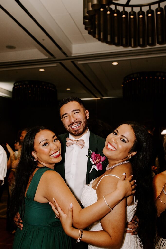 A bride and two members of her wedding party smile at the camera while dancing together