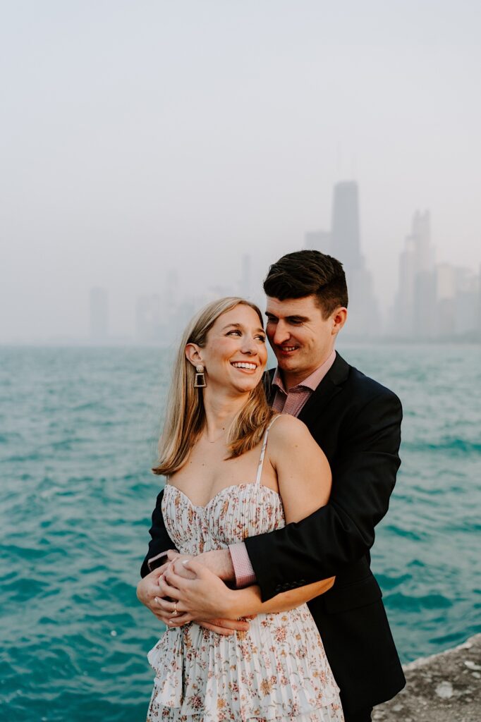 A woman looks over her shoulder and smiles to a man who is hugging her from behind and smiling at her as they stand in front of Lake Michigan and the Chicago skyline