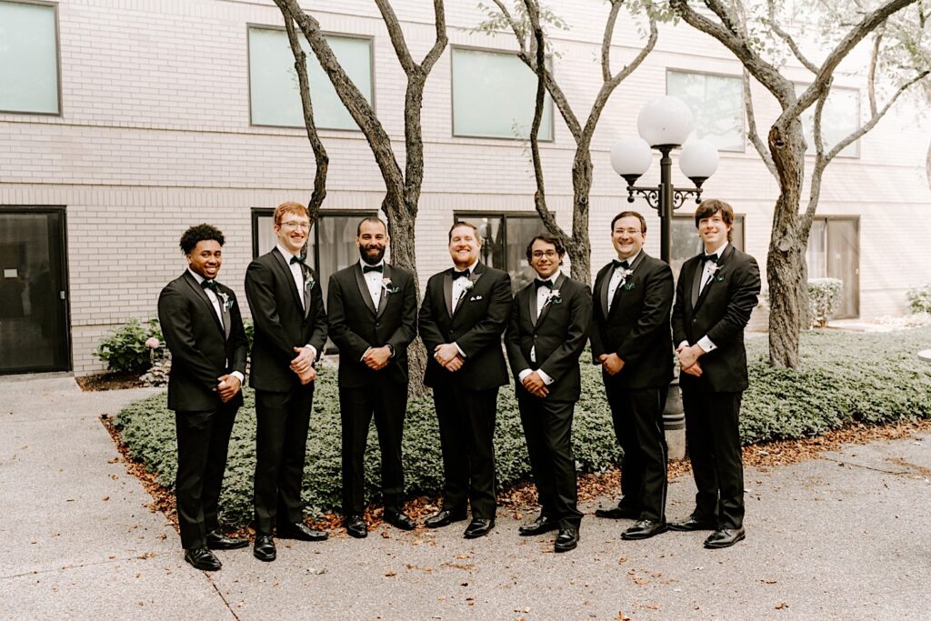 A groom and his groomsmen pose with one another and smile at the camera while standing outside of a building with trees behind them