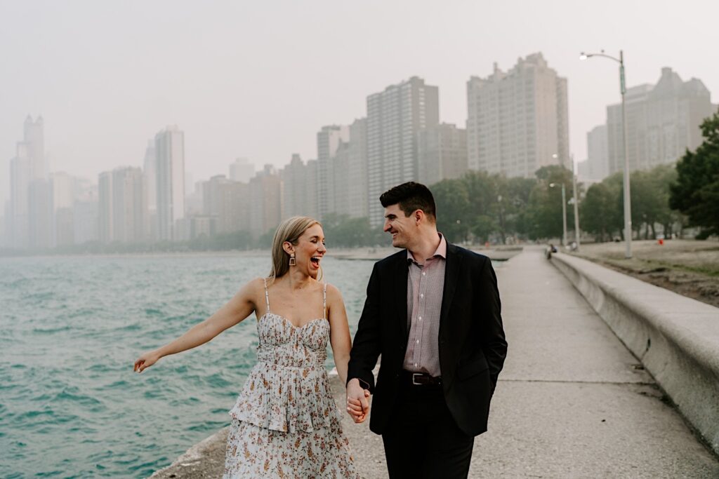 A man and woman walk hand in hand along Lake Michigan and smile at one another during their engagement session with a hazy Chicago skyline behind them