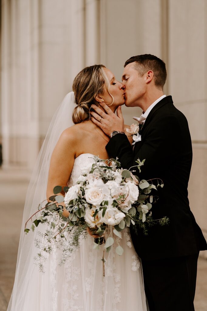 A bride and groom kiss one another while inside a building, the bride is holding her bouquet while the groom holds her face