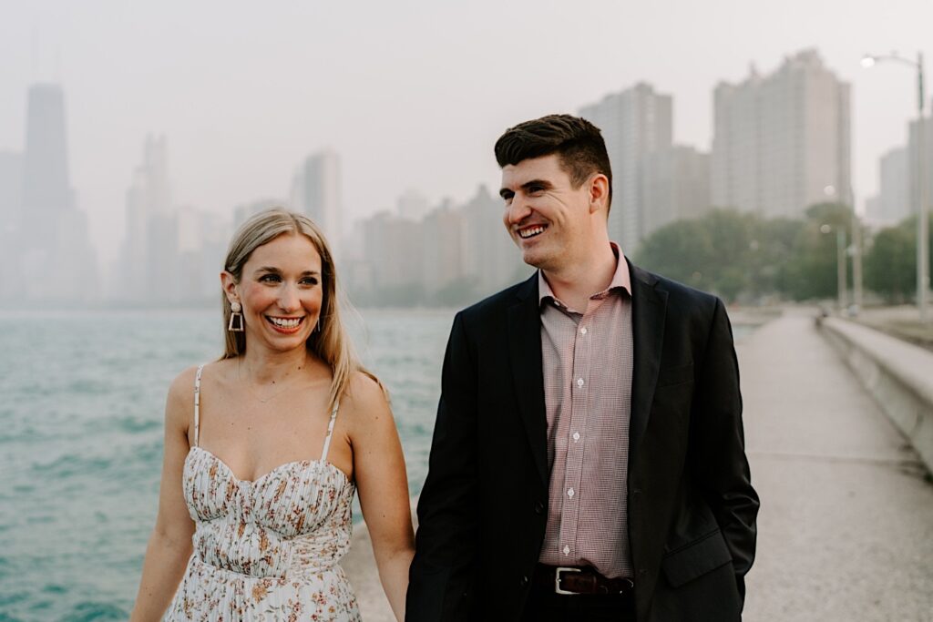A man and woman smile while walking and holding hands along Lake Michigan  during their engagement session with a hazy Chicago skyline behind them