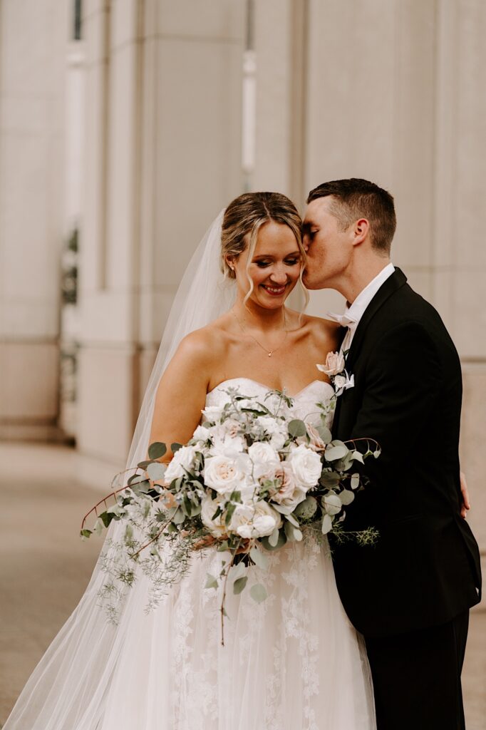 A bride smiles down at her bouquet as the groom kisses her on the cheek while the two stand inside a building