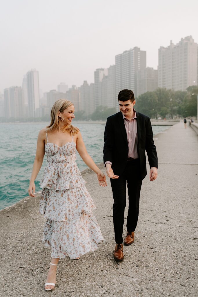A woman looks over her shoulder and reaches her hand out behind her towards a man who is reaching to hold her hand as they both smile while walking along Lake Michigan