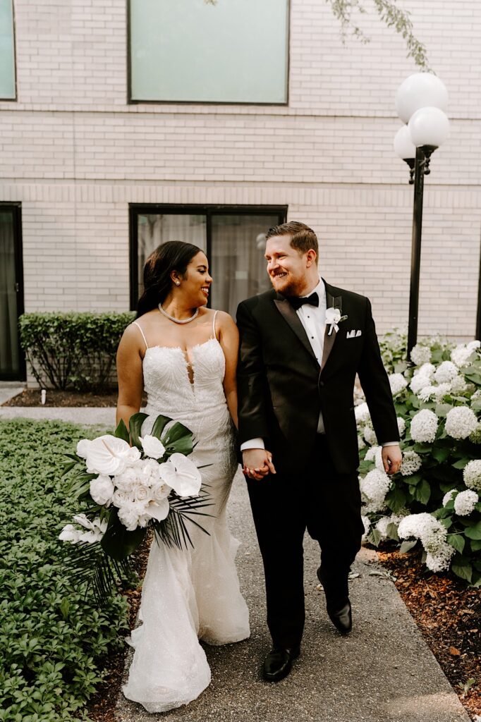 A bride and groom smile at one another and walk hand in hand on a path outside of a building towards the camera