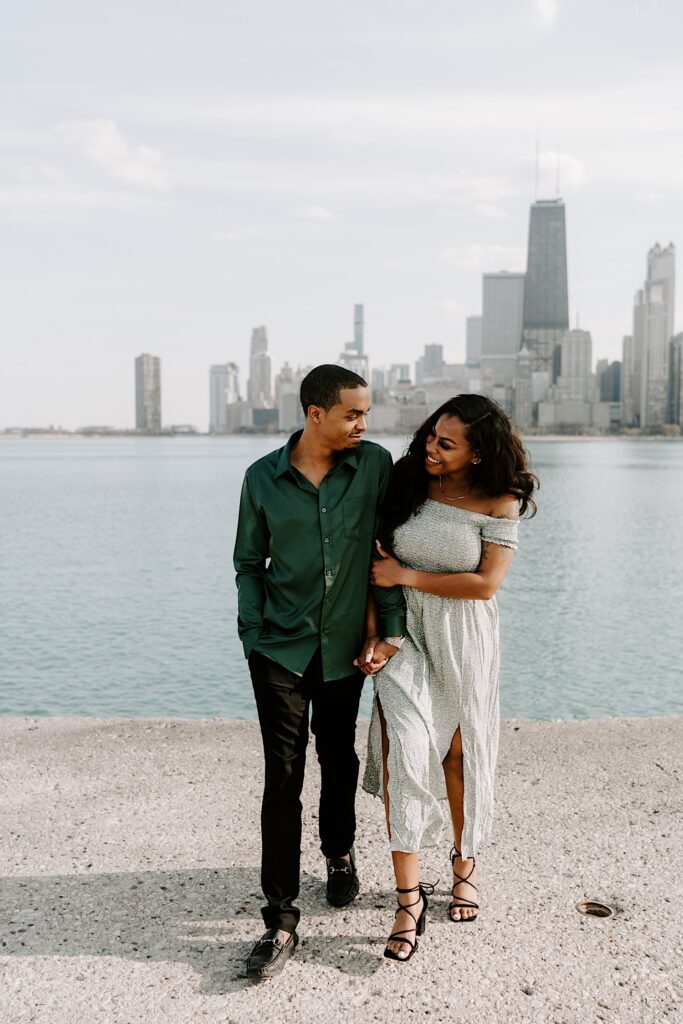 A man and woman walk with one another while smiling at each other at North Avenue Beach with the Chicago skyline and Lake Michigan behind them