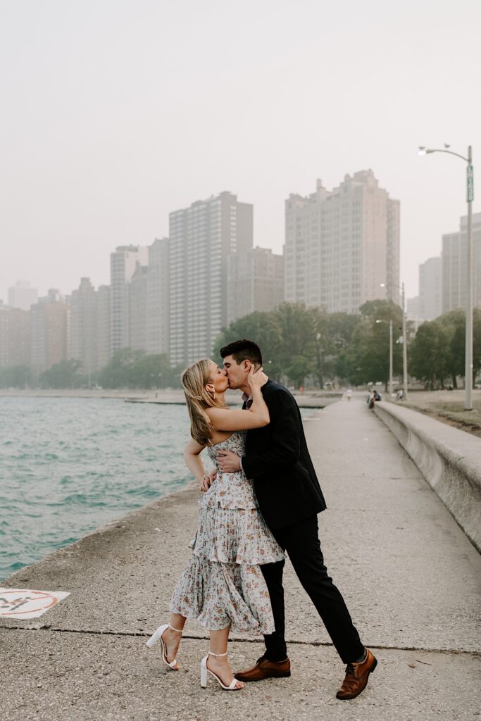 A man and woman kiss one another and embrace while standing next to Lake Michigan and with the Chicago skyline behind them