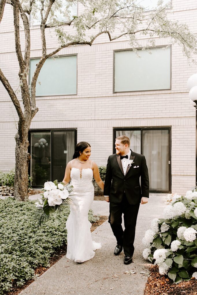 A bride and groom smile at one another and walk hand in hand on a path outside of a building towards the camera
