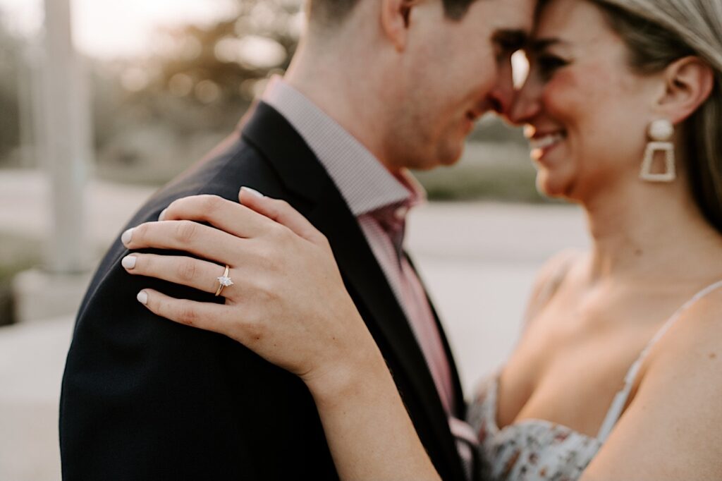 A woman's hand with an engagement ring rests on a man's shoulder as they both smile at one another in the background with their noses touching