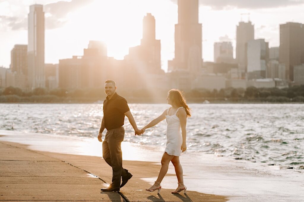 A man and woman smile while walking and holding hands in front of Lake Michigan with the sunset and the Chicago skyline behind them during their engagement photos