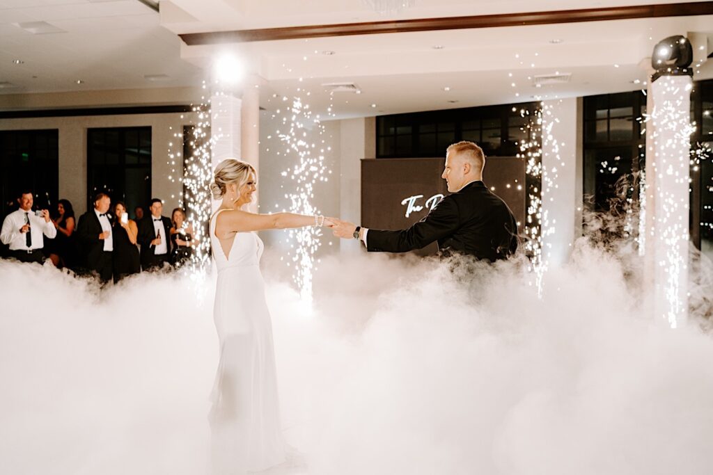 A bride and groom hold hands while sharing their first dance during their indoor reception, around them is smoke from a smoke machine and 4 vertical pillars of sparks