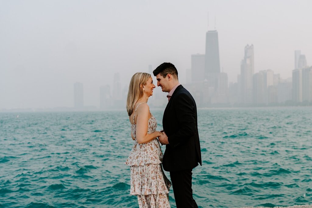 A man and woman smile at one another while holding hands and facing each other during their engagement session with Lake Michigan and the hazy Chicago skyline behind them