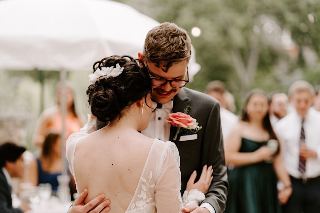 A groom smiles as the bride puts her head on his chest while they share their first dance during their outdoor reception while guests watch in the background