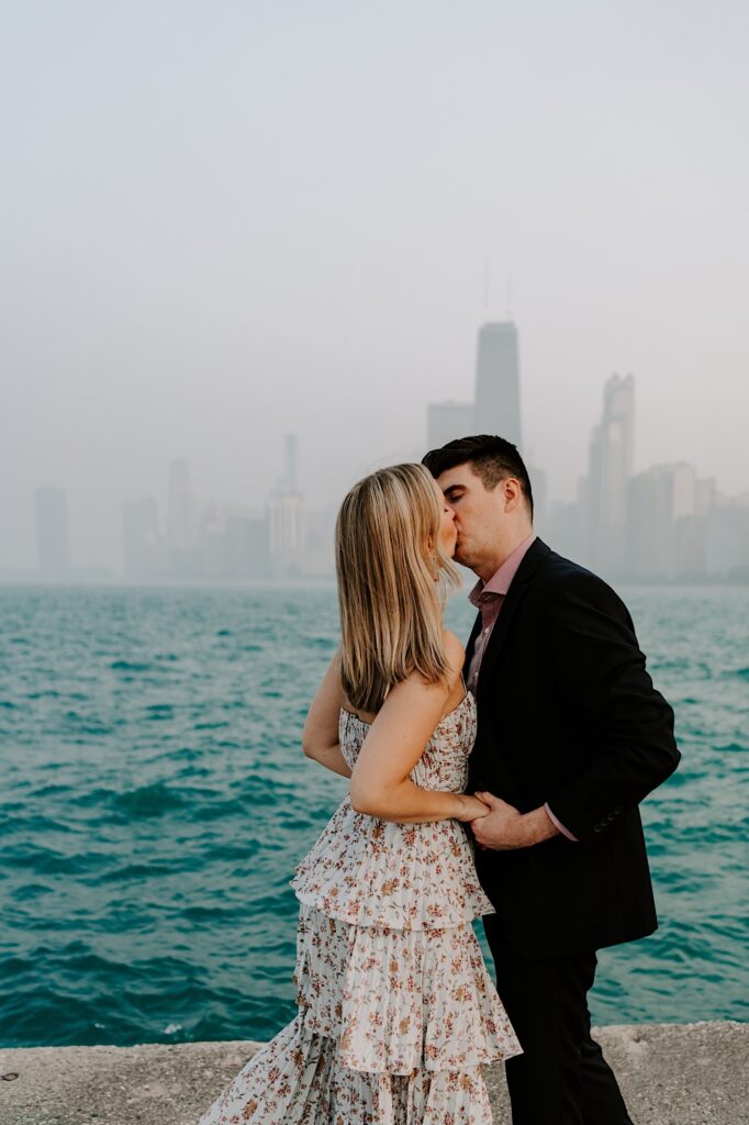 A man and woman kiss one another while holding hands with Lake Michigan and the Chicago skyline behind them