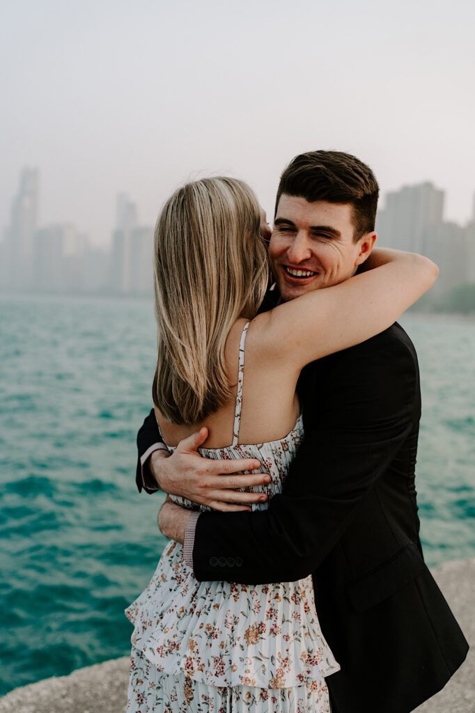 A man smiles at the camera while hugging a woman with her back to the camera with Lake Michigan and the Chicago skyline behind them