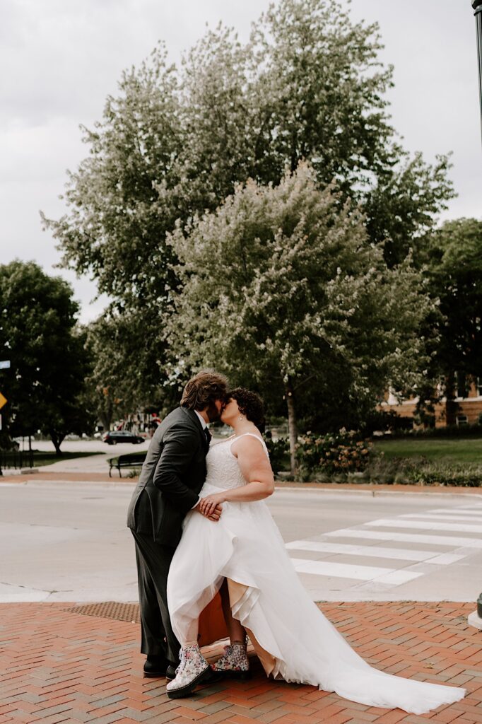A bride and groom stand on a brick sidewalk and kiss one another with an empty street and a park with trees behind them