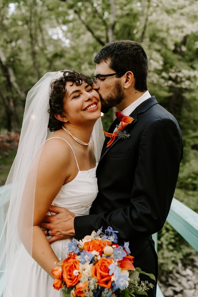 A bride smiles and closes her eyes while the groom hugs her and kisses her cheek while they stand outside on a bridge with trees in the background